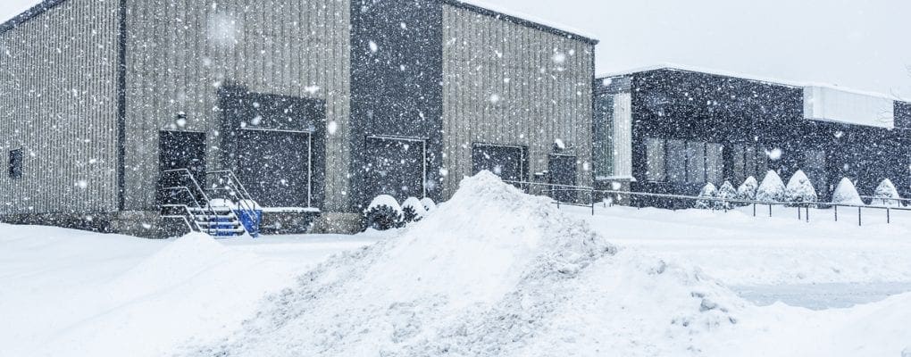 Industrial warehouse loading docks and attached commercial office building behind a heap of plowed and drifting snow at the edge of the parking lot during a blinding blizzard snow storm.