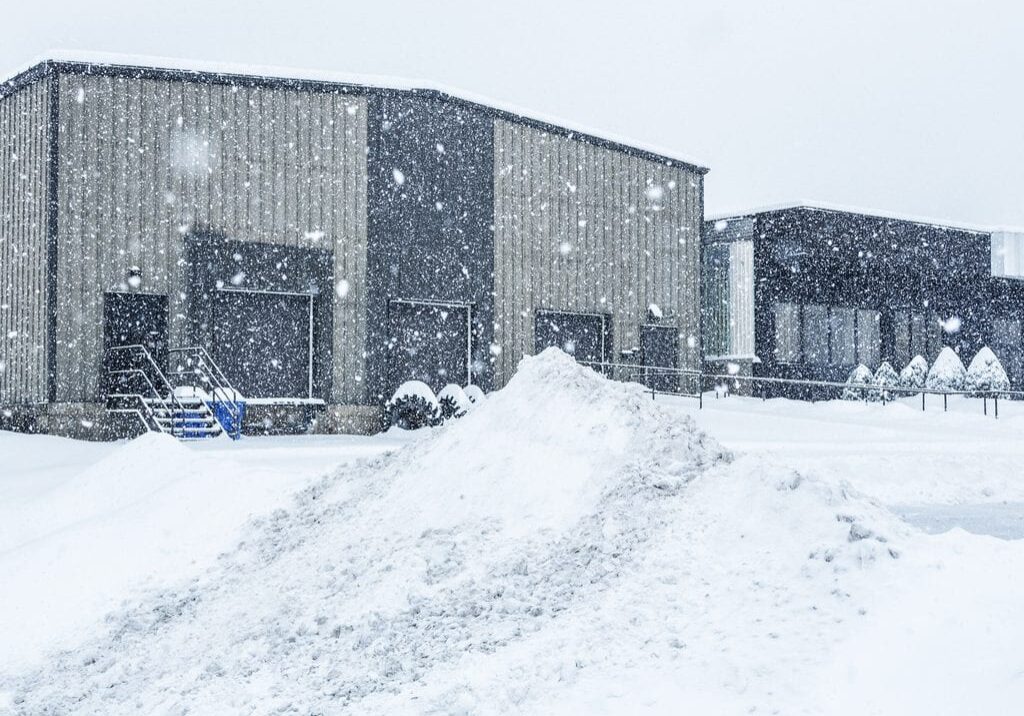 Industrial warehouse loading docks and attached commercial office building behind a heap of plowed and drifting snow at the edge of the parking lot during a blinding blizzard snow storm.